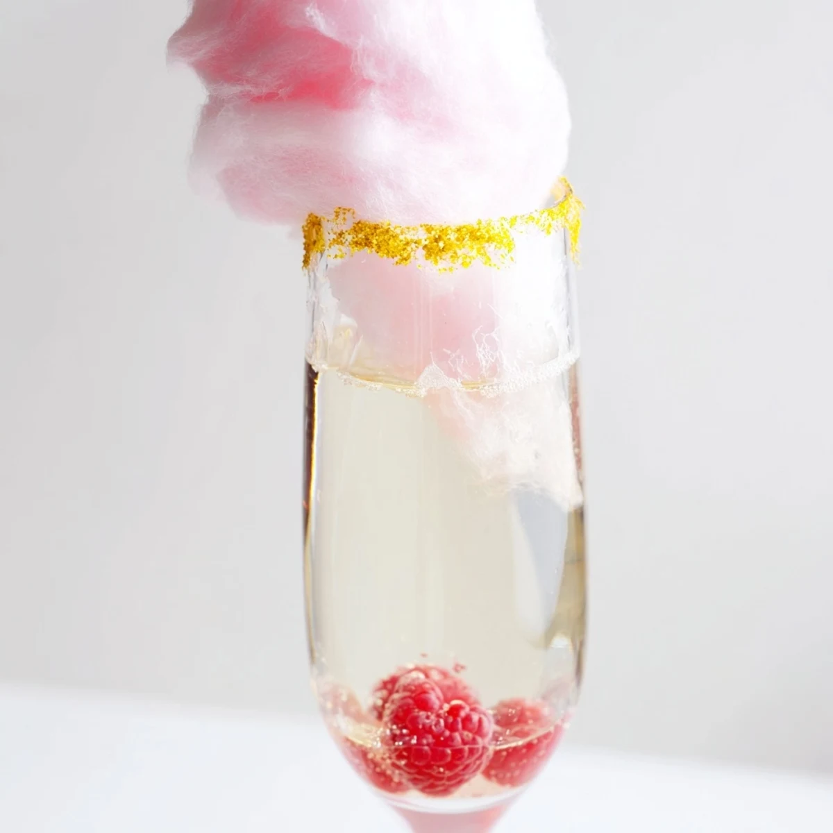 Glossy pink mocktail topped with cotton candy and edible glitter, served in a stemmed flute on a marble surface for a festive, bubbly party centerpiece.