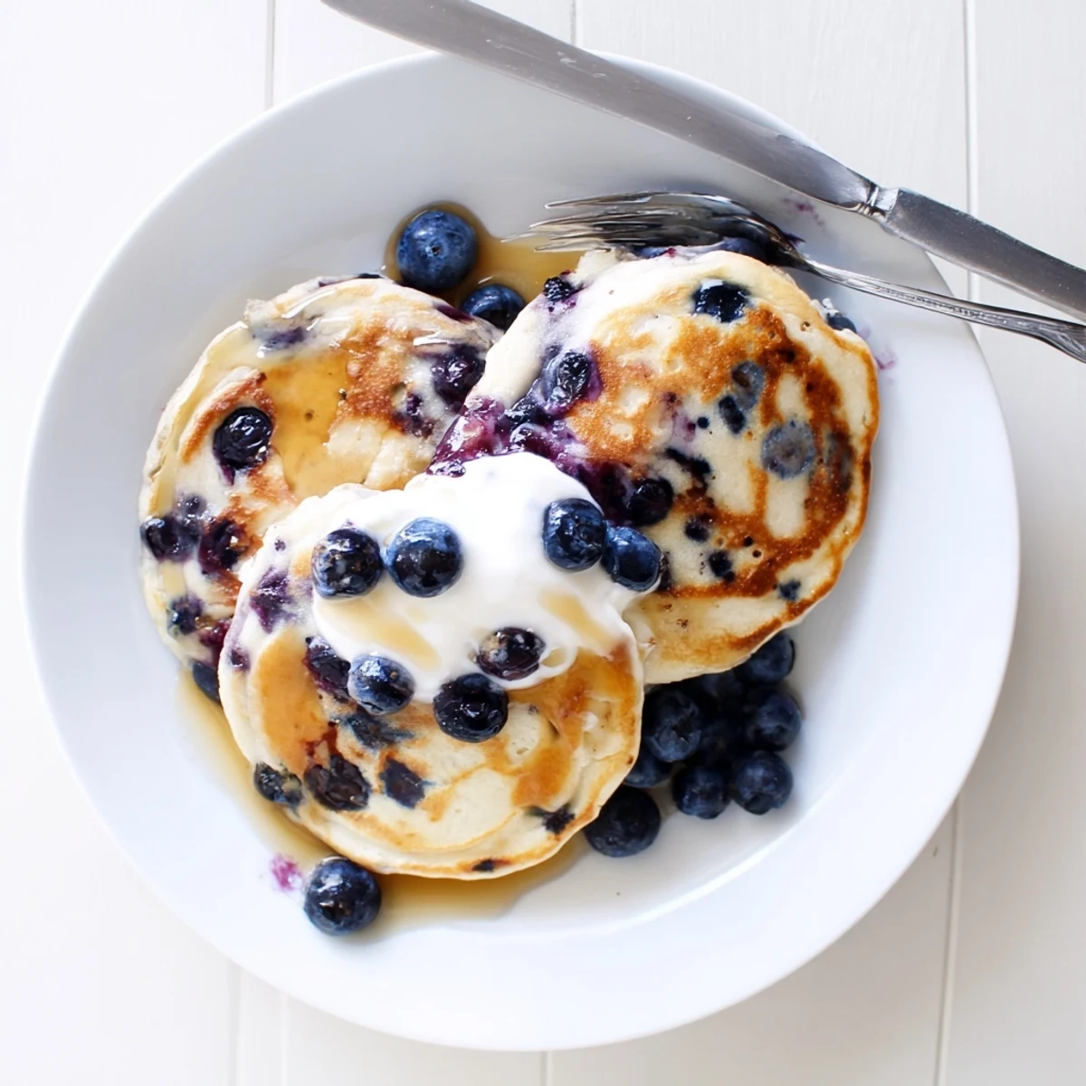 A close-up of fluffy Greek Yogurt Blueberry Pancakes dripping with maple syrup and topped with extra blueberries.
